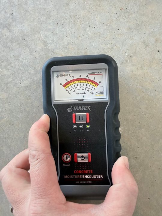 Hand holding a Tramex concrete moisture meter on a bare concrete slab